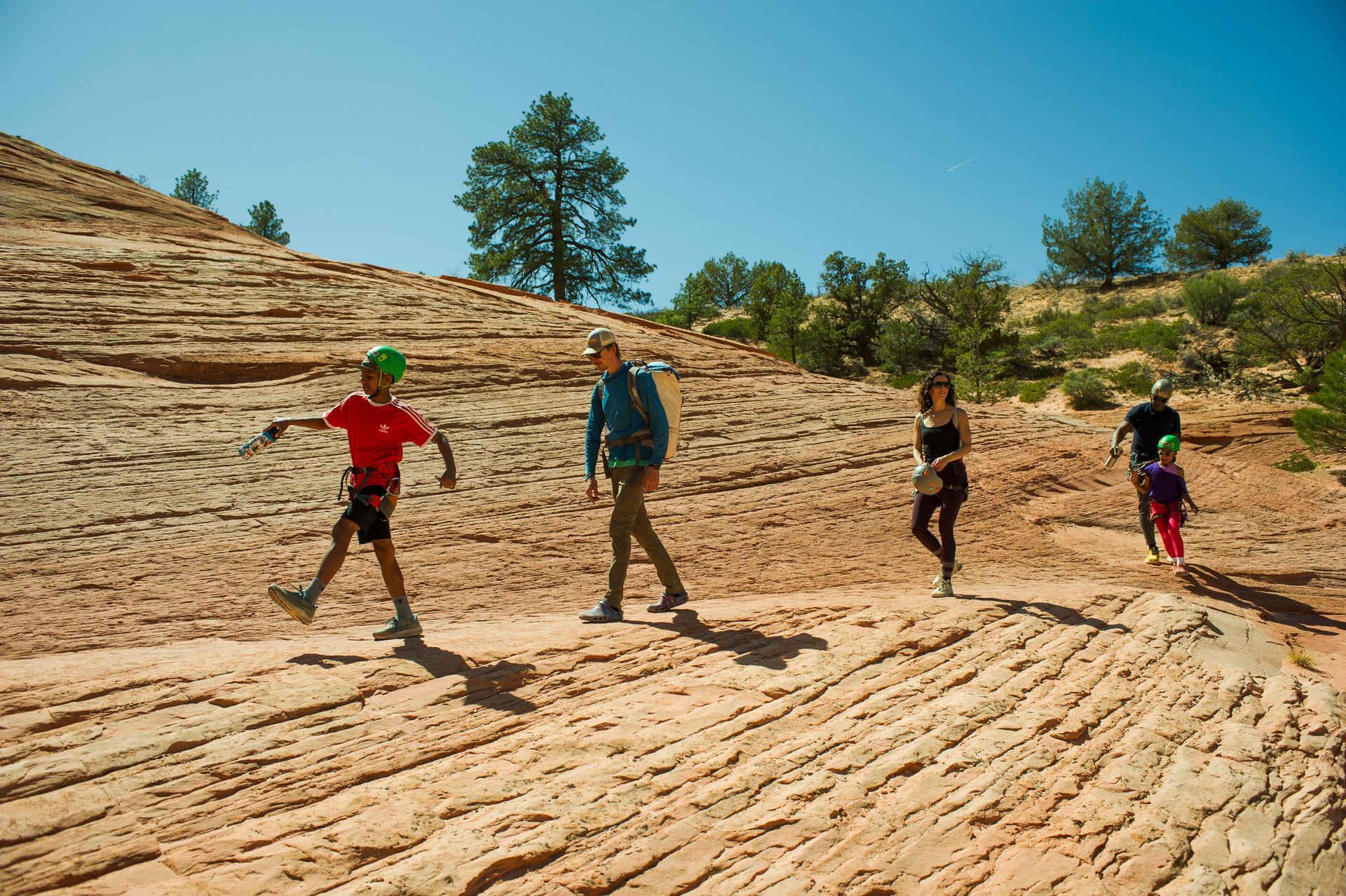 A group of people wearing helmets and backpacks hike along a textured, sloping sandstone trail under a clear blue sky.