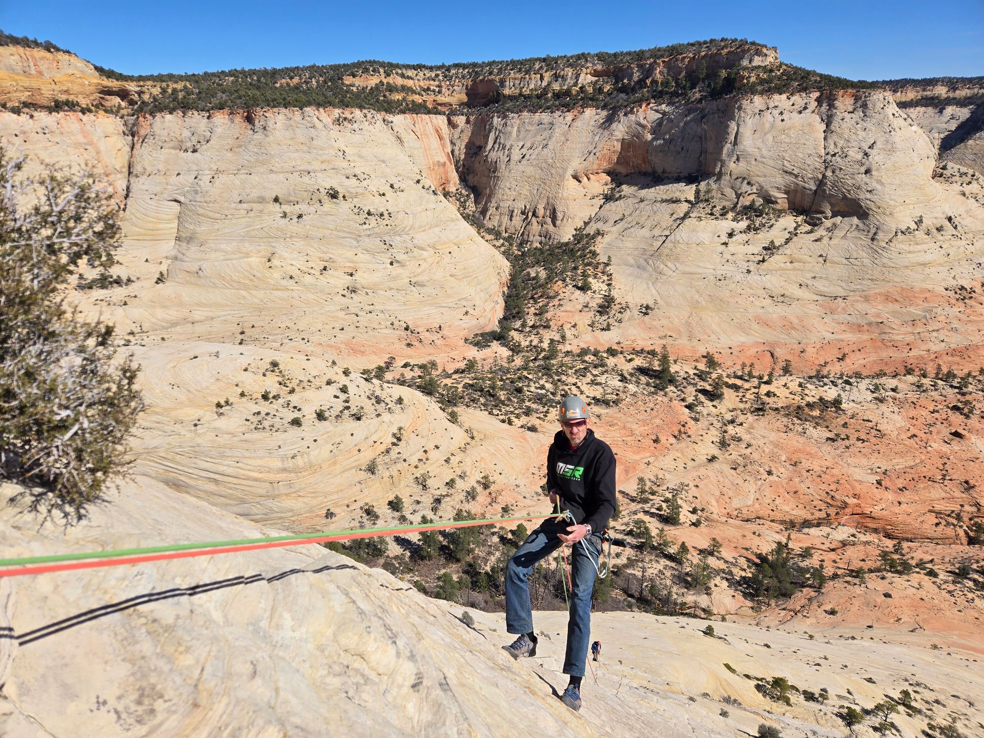 A person in a black jacket and jeans prepares to rappel down a light-colored sandstone cliff in a desert canyon.
