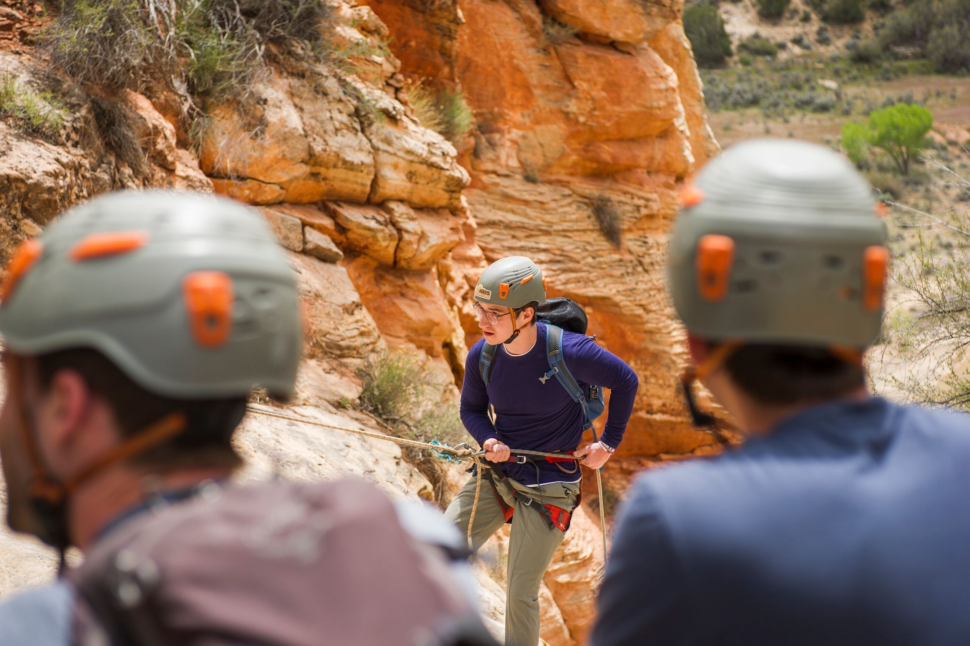 Climbers in helmets and harnesses on a red rock canyon wall, with one instructor in front