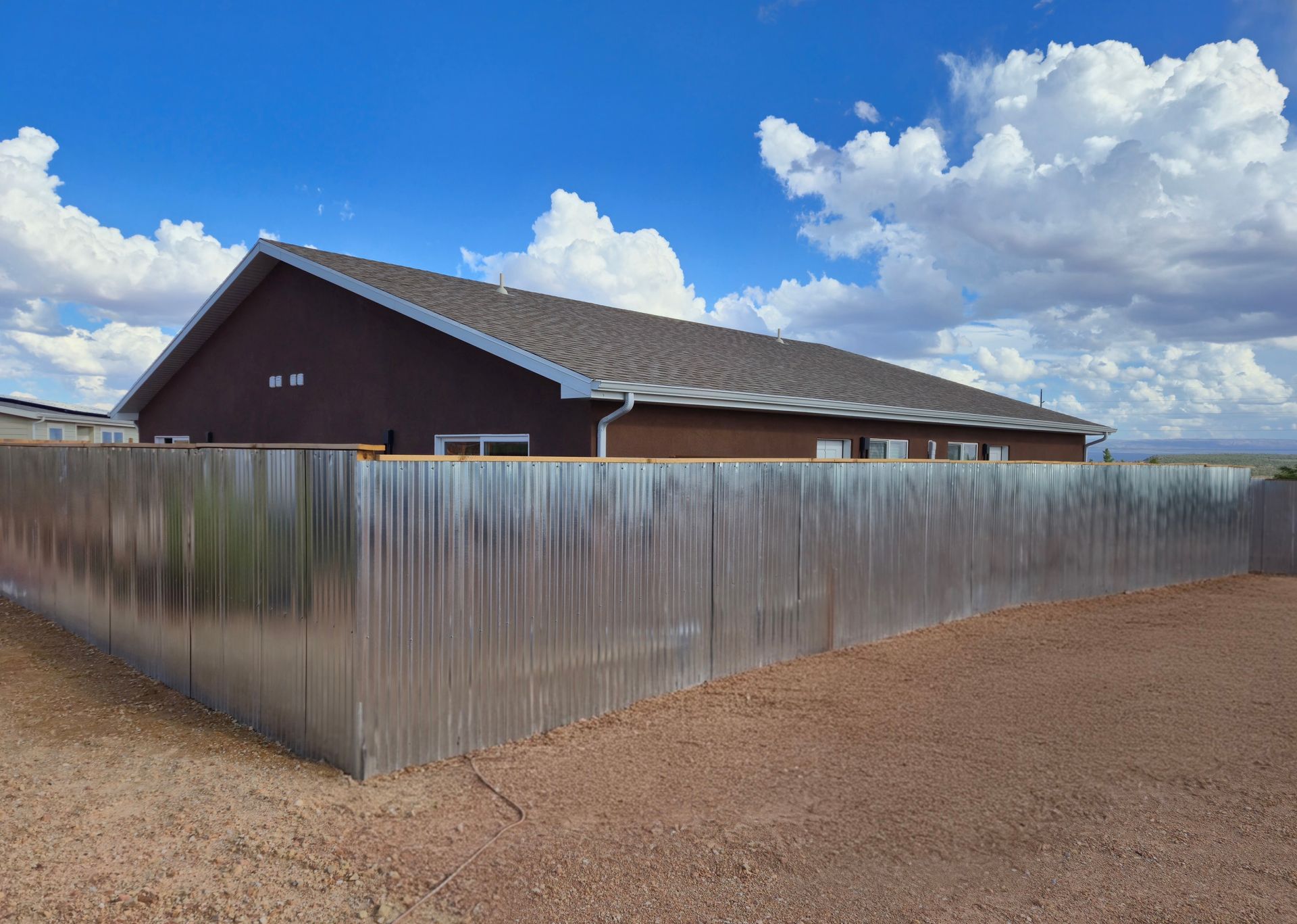 Metal fence around a brown house under a bright blue sky with clouds.