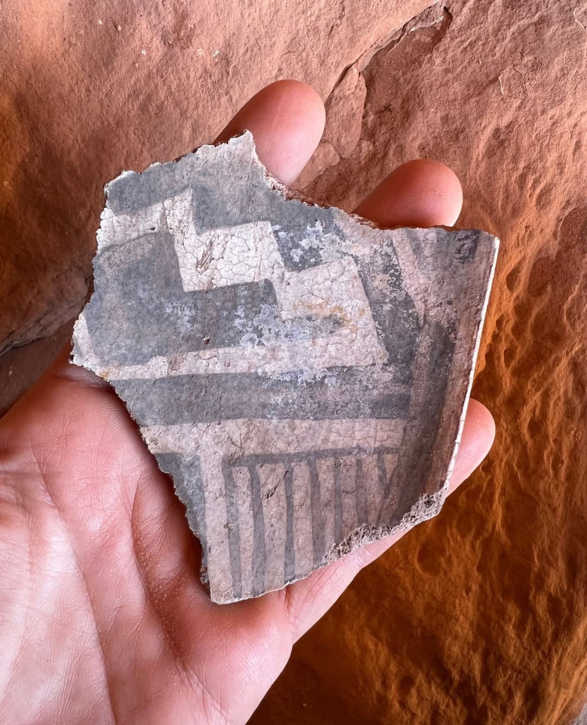 Hand holding a fragment of gray and white ancient pottery, with geometric designs, against a red rock background.