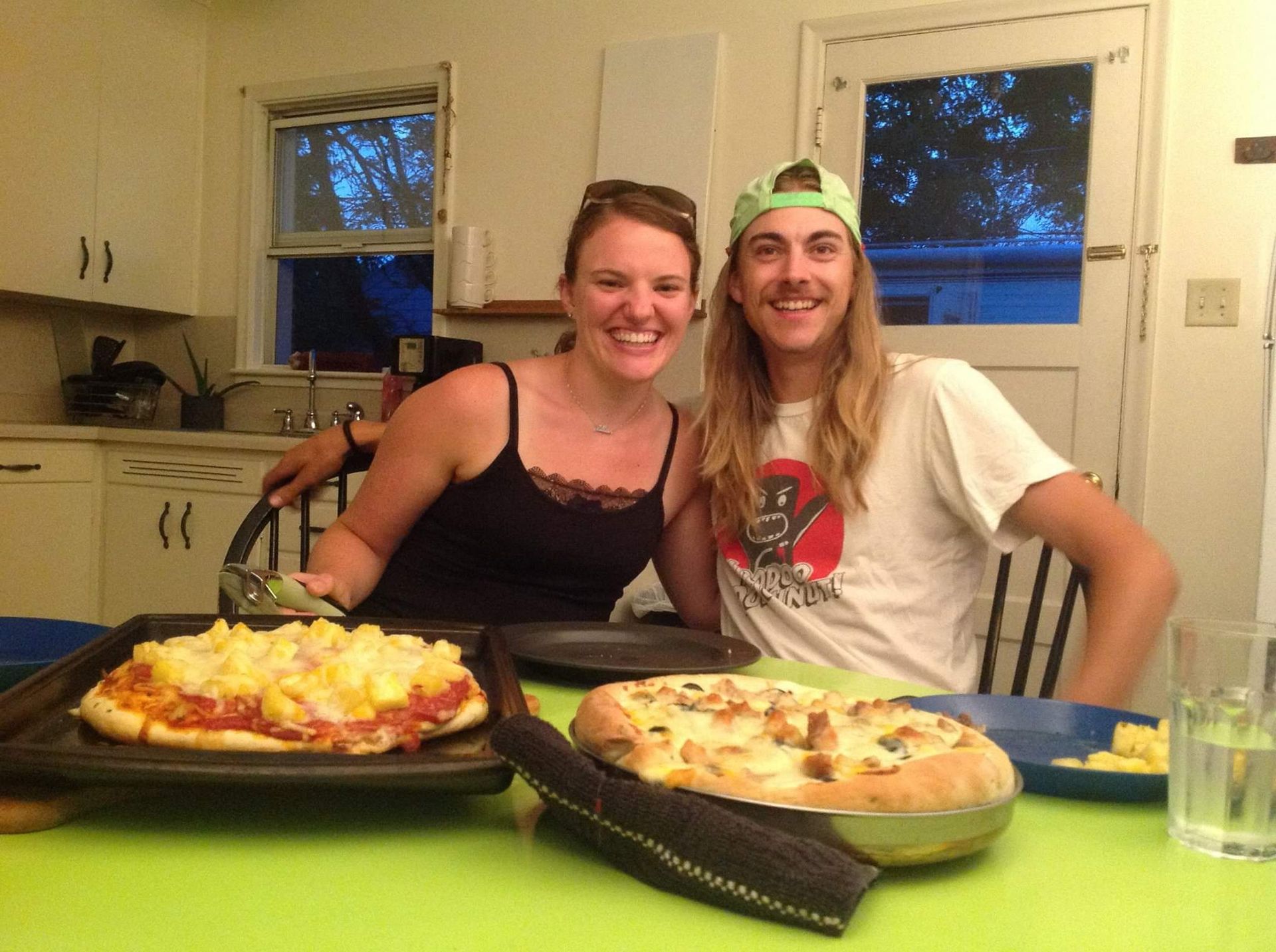 Two people smile at the camera, pizza on a table in a kitchen.