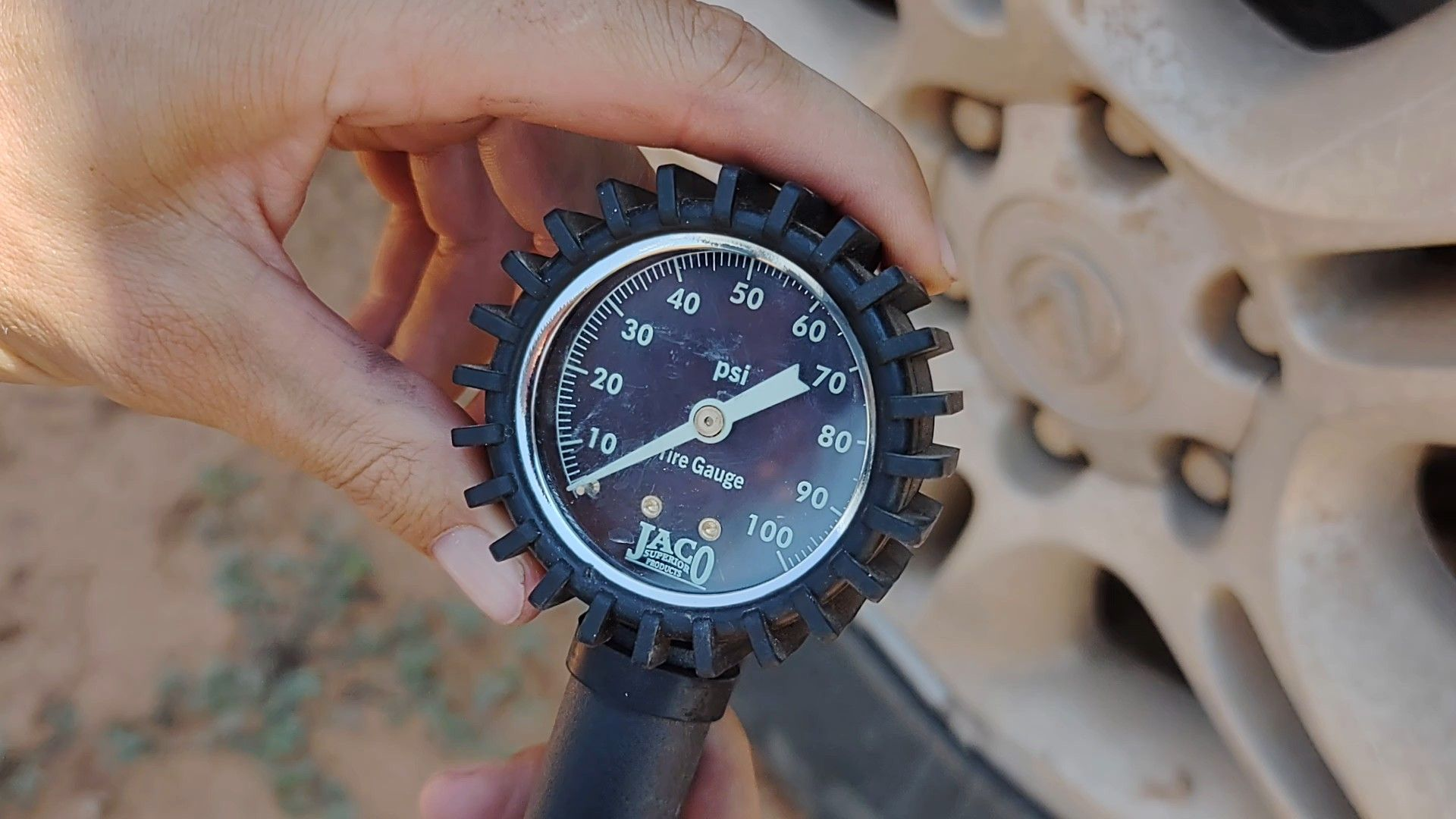 Hand holding a tire pressure gauge; reading approximately 65 PSI near a car tire.