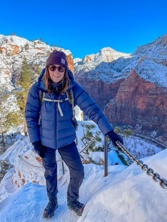 Woman in blue jacket on snowy mountain trail, holding chain. Zion National Park with blue sky.