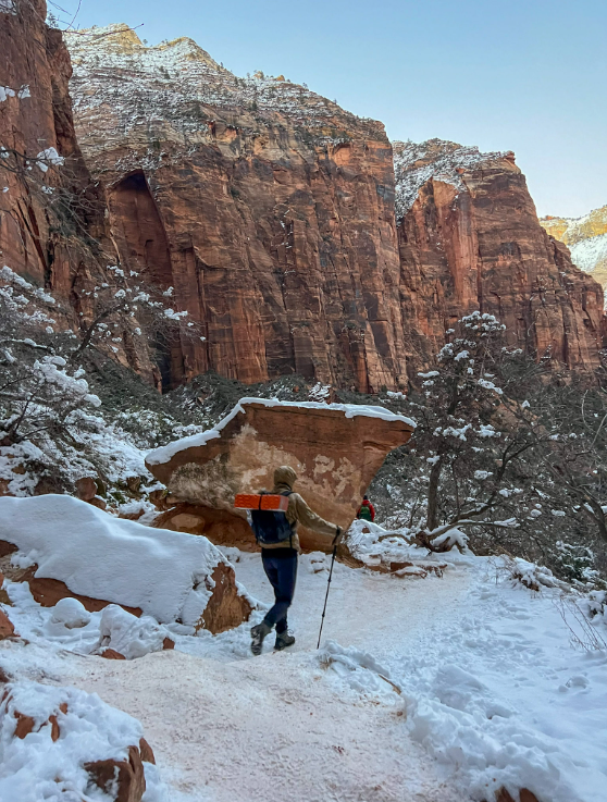 Person hikes snowy trail in Zion National Park, red rock cliffs in background.