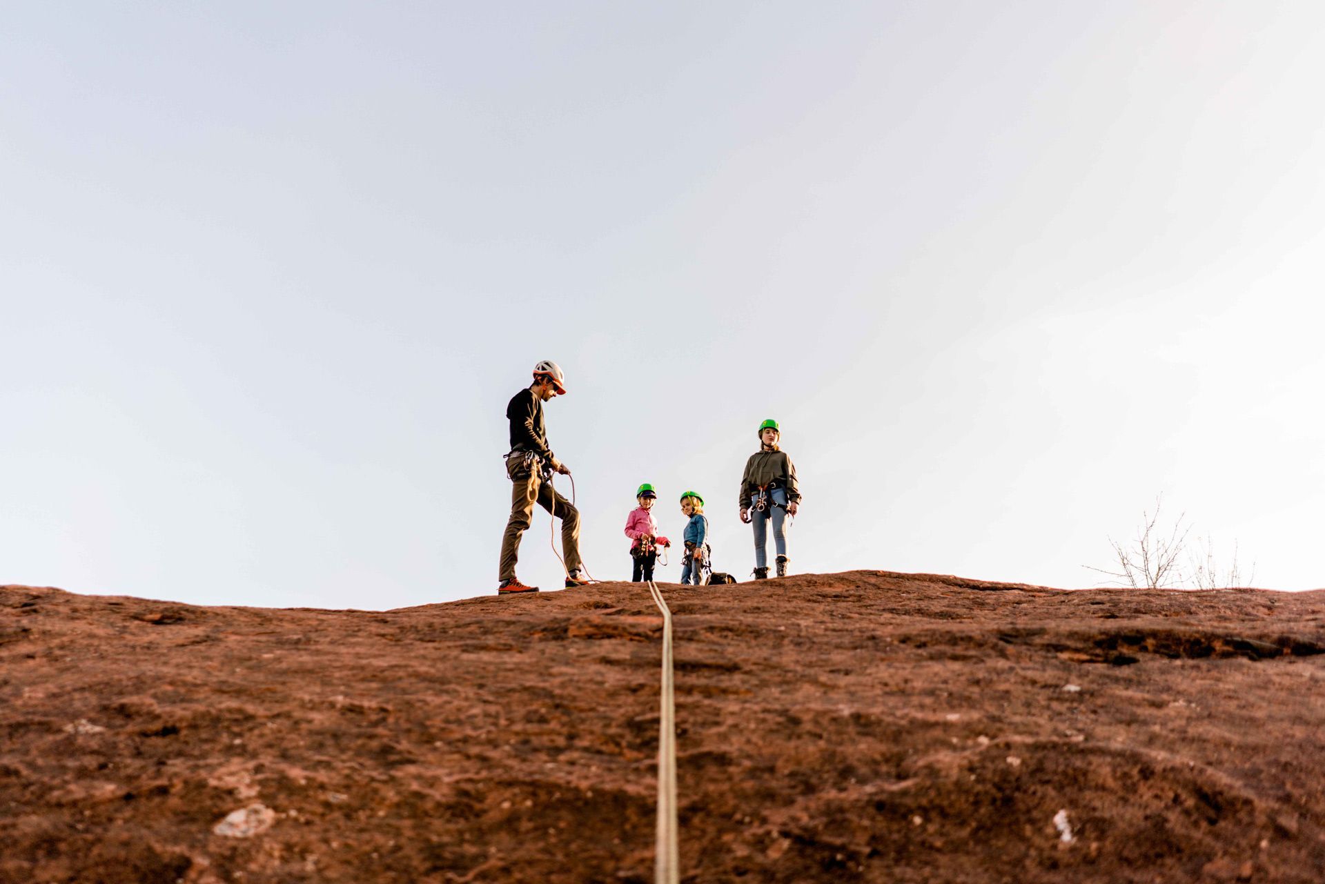 People rappelling down a red rock cliff. A man guides two children, roped in, on a clear day.