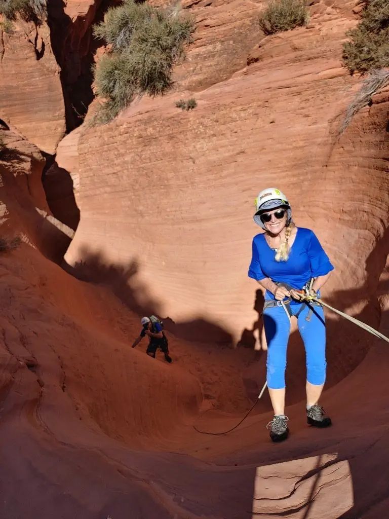 Woman rappels down a red rock canyon, with another person visible below. She wears a helmet and blue clothes, smiling.