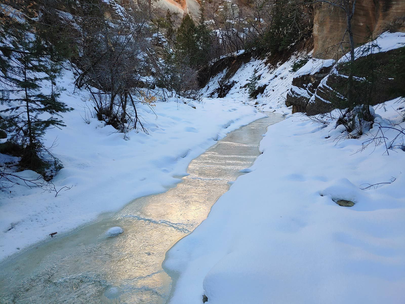 A snowy canyon with a partially frozen stream winding through it. Sunlight glints off the icy water.