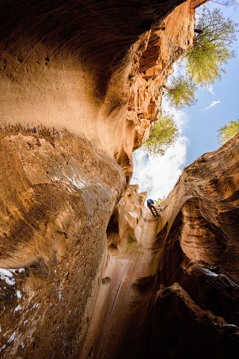 A cyclist rides down a narrow canyon trail, viewed from below. Brown rock walls surround the path; blue sky.
