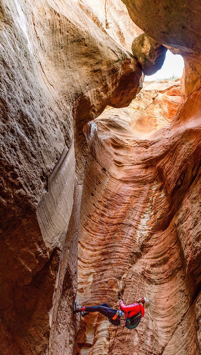 A person rappels down a narrow, orange-and-tan canyon, with sunlight streaming from above.