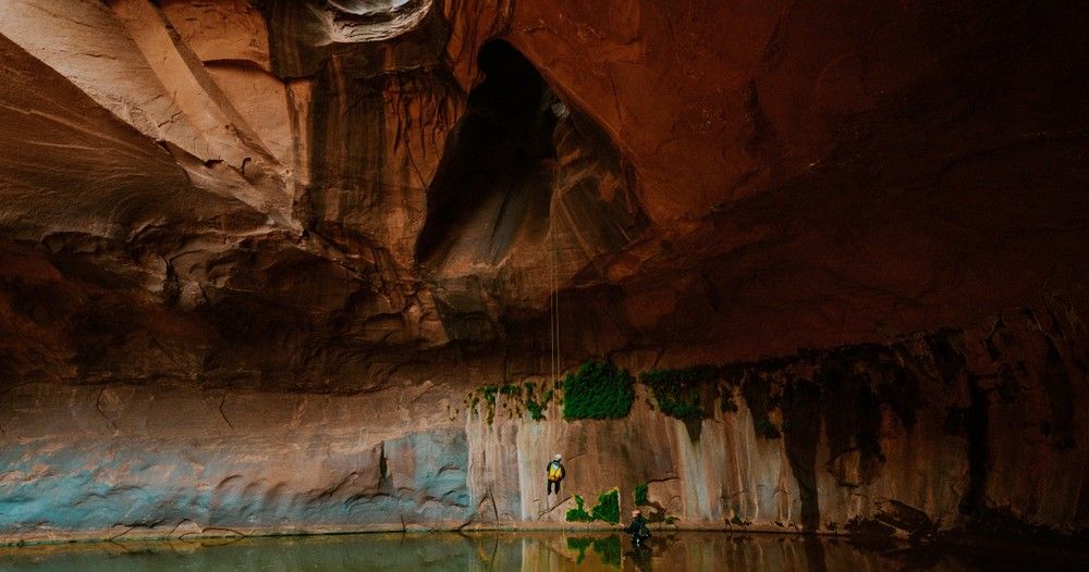 Cave interior with pool of water reflecting the walls. A person stands near a wall with green growth.