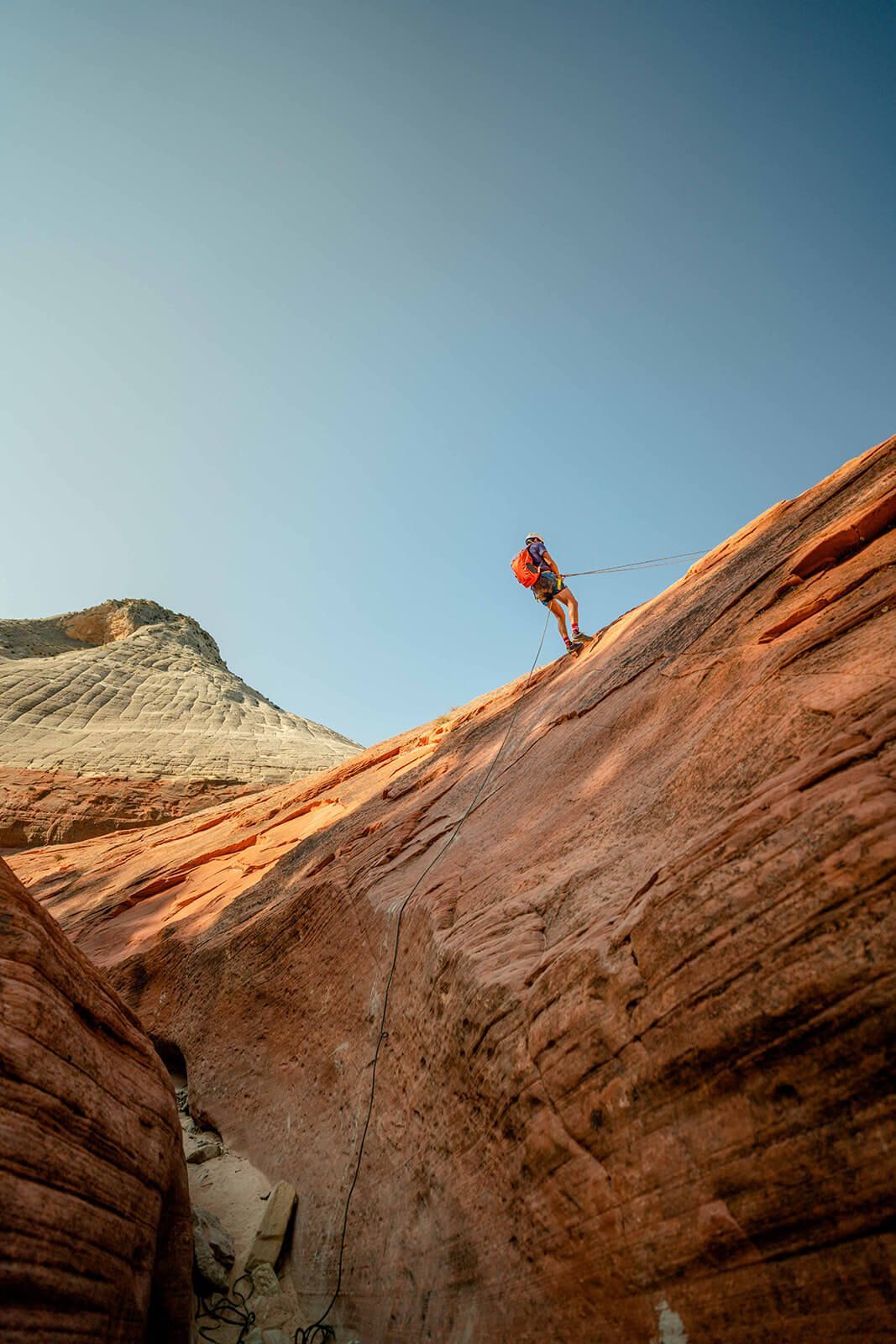 Rock climber rappelling down a steep, red rock face under a clear blue sky.