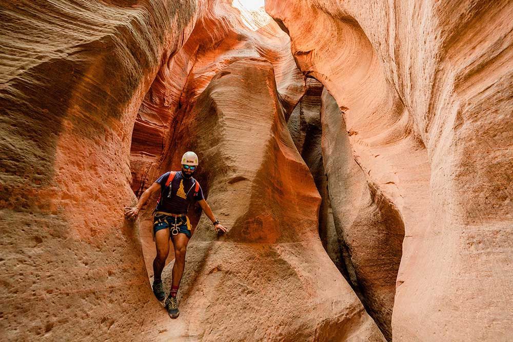 A person navigates a narrow, orange sandstone canyon, wearing a helmet and gear, reaching out to walls.