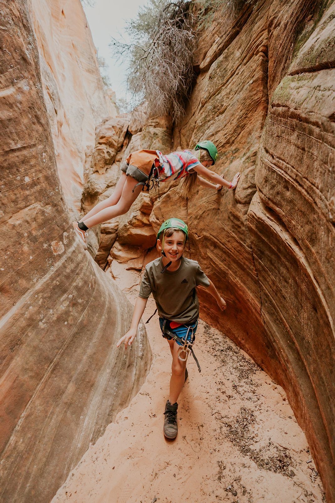 Two kids canyoneering in a red rock slot canyon; one climbing, the other smiling.