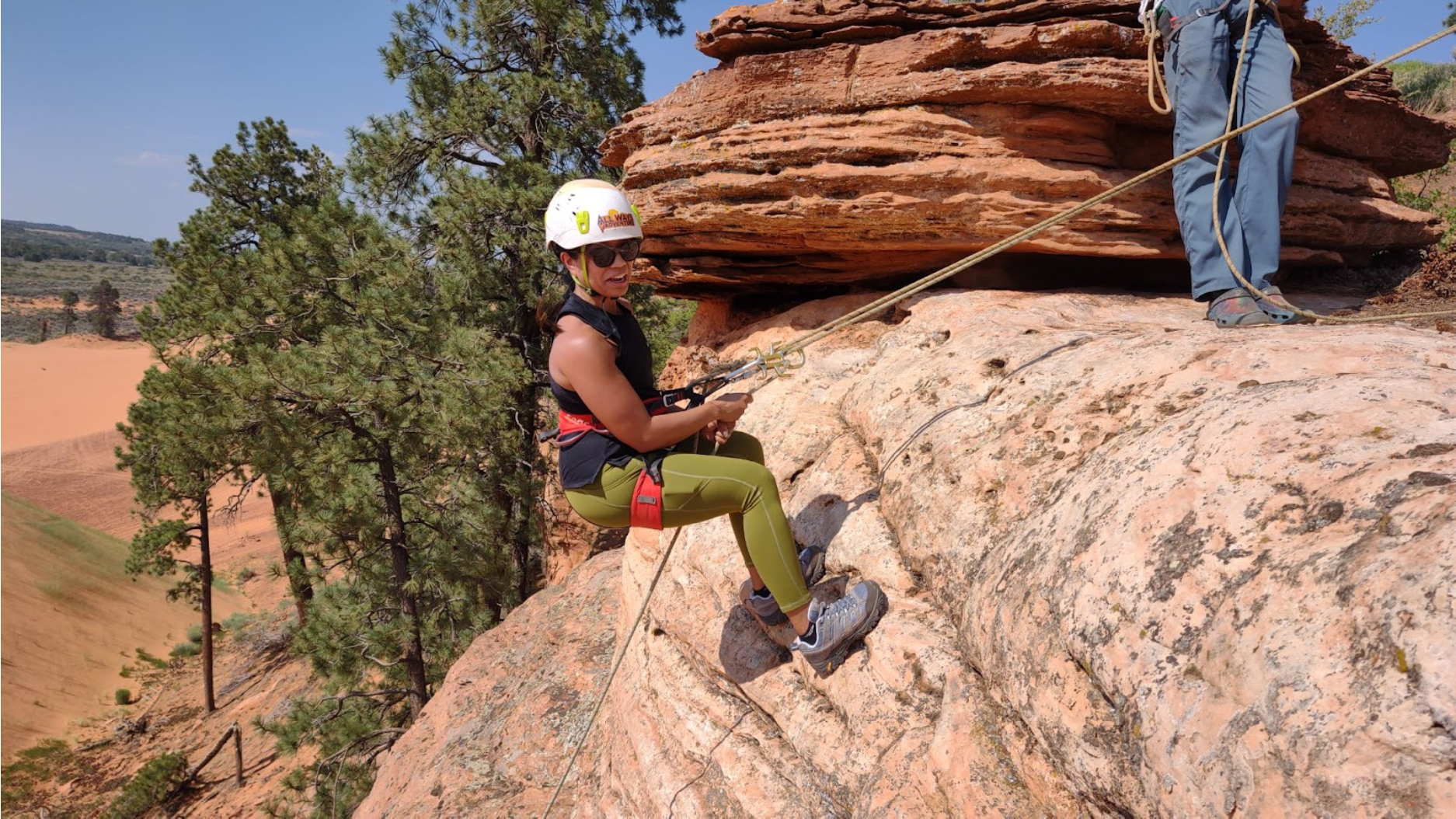 Woman rappels down a sandstone cliff, wearing a helmet and safety harness. Person above anchors the rope.