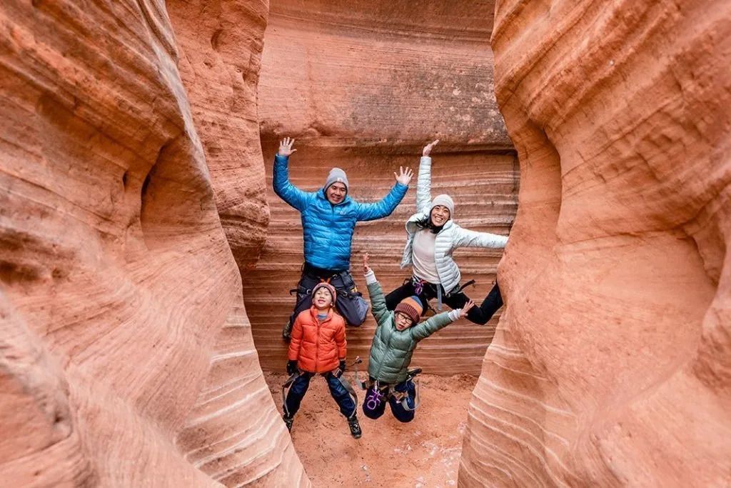 Family in canyon, arms raised, smiling. Red rock walls, blue and orange jackets.