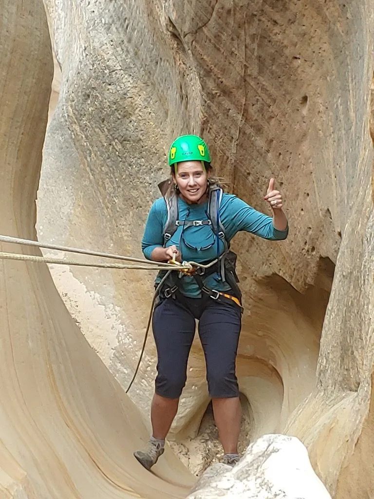Woman rappelling in a sandstone canyon, wearing a green helmet, blue shirt, and giving a thumbs-up.