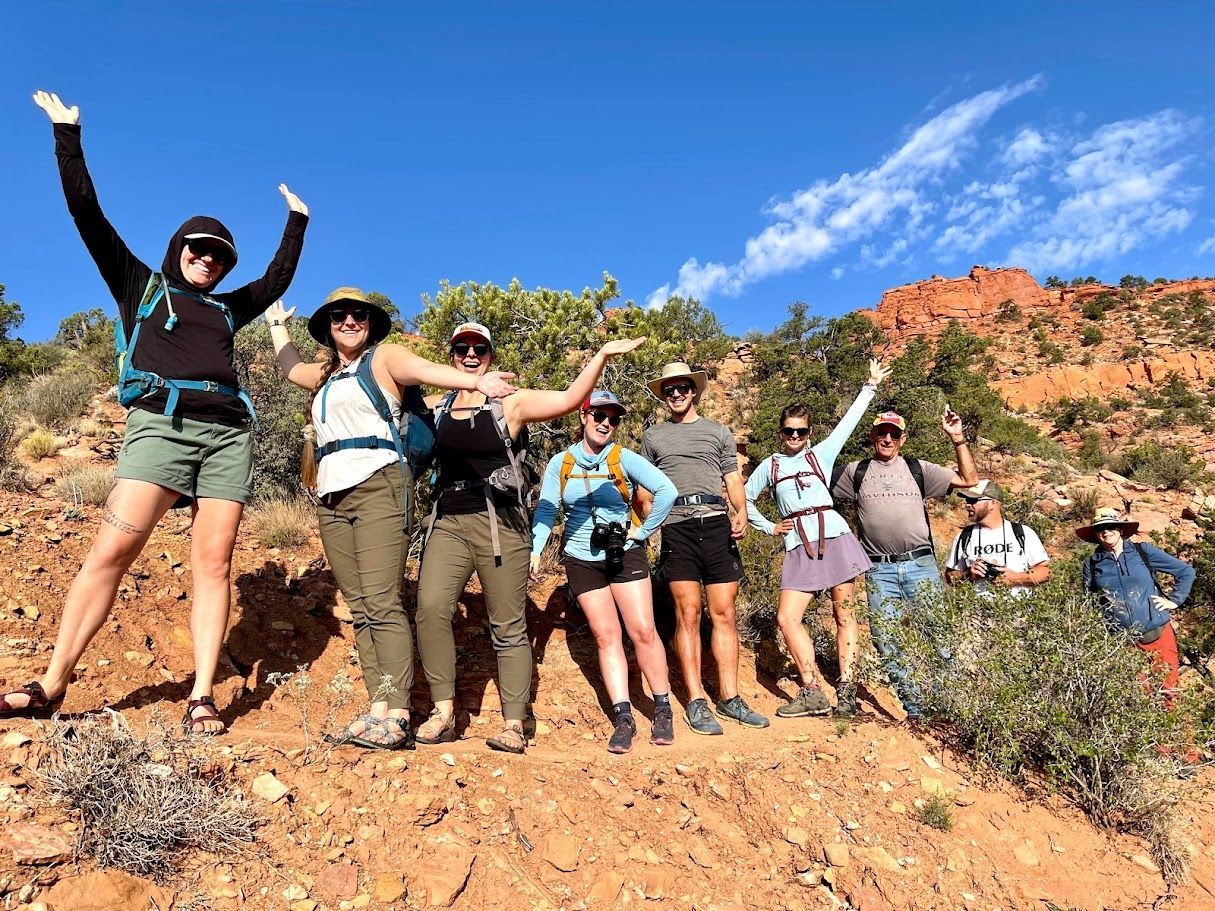 Group of hikers with raised arms on a red dirt trail against a blue sky and red rock formations.