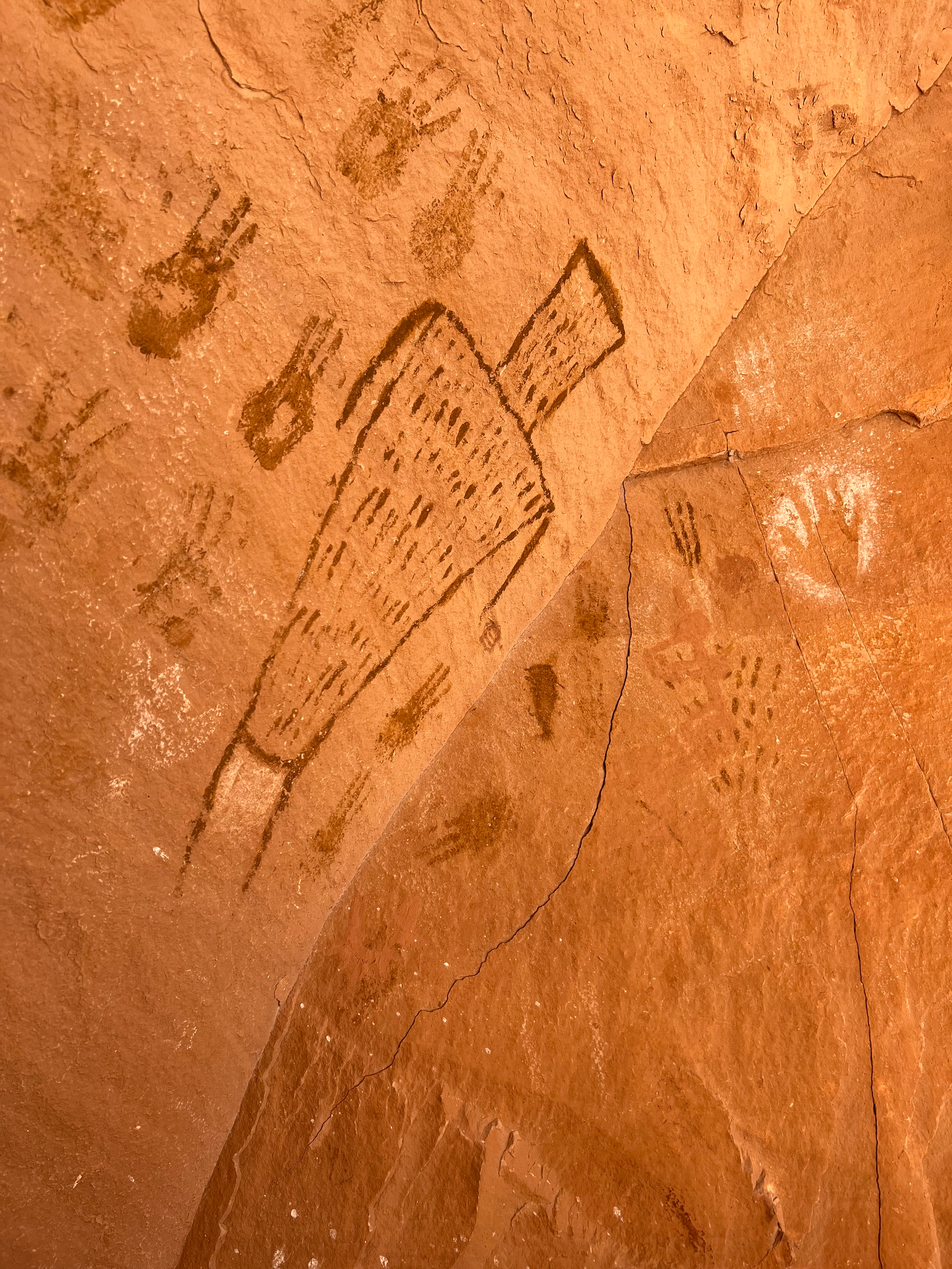 Cave painting of a person with a large headdress. Handprints surround it in a tan cave setting.