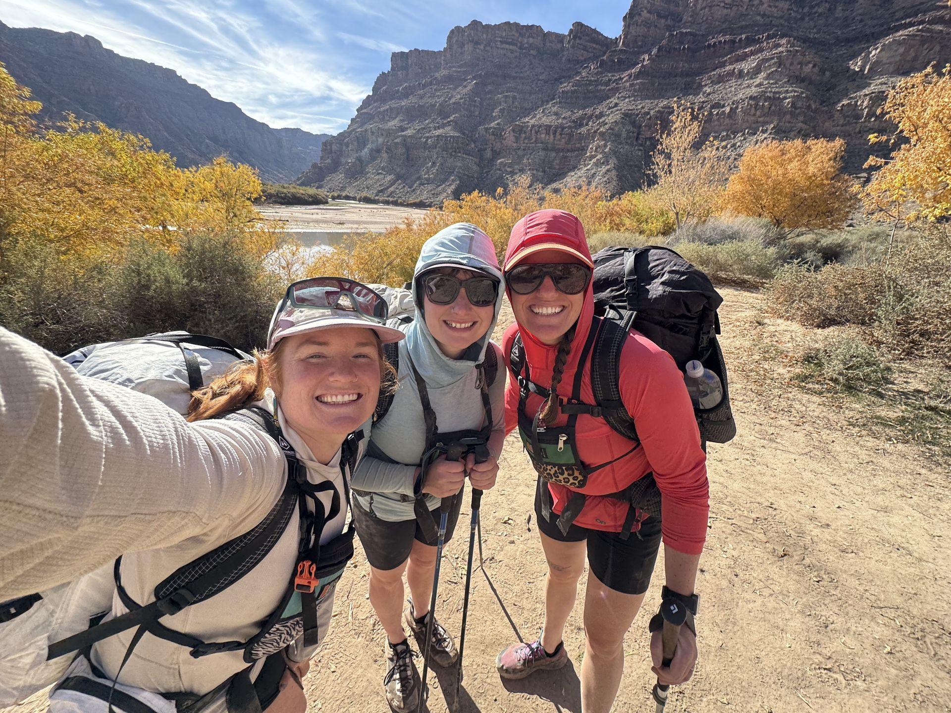 Three hikers pose for selfie on a trail in a canyon with fall foliage; sunny day.