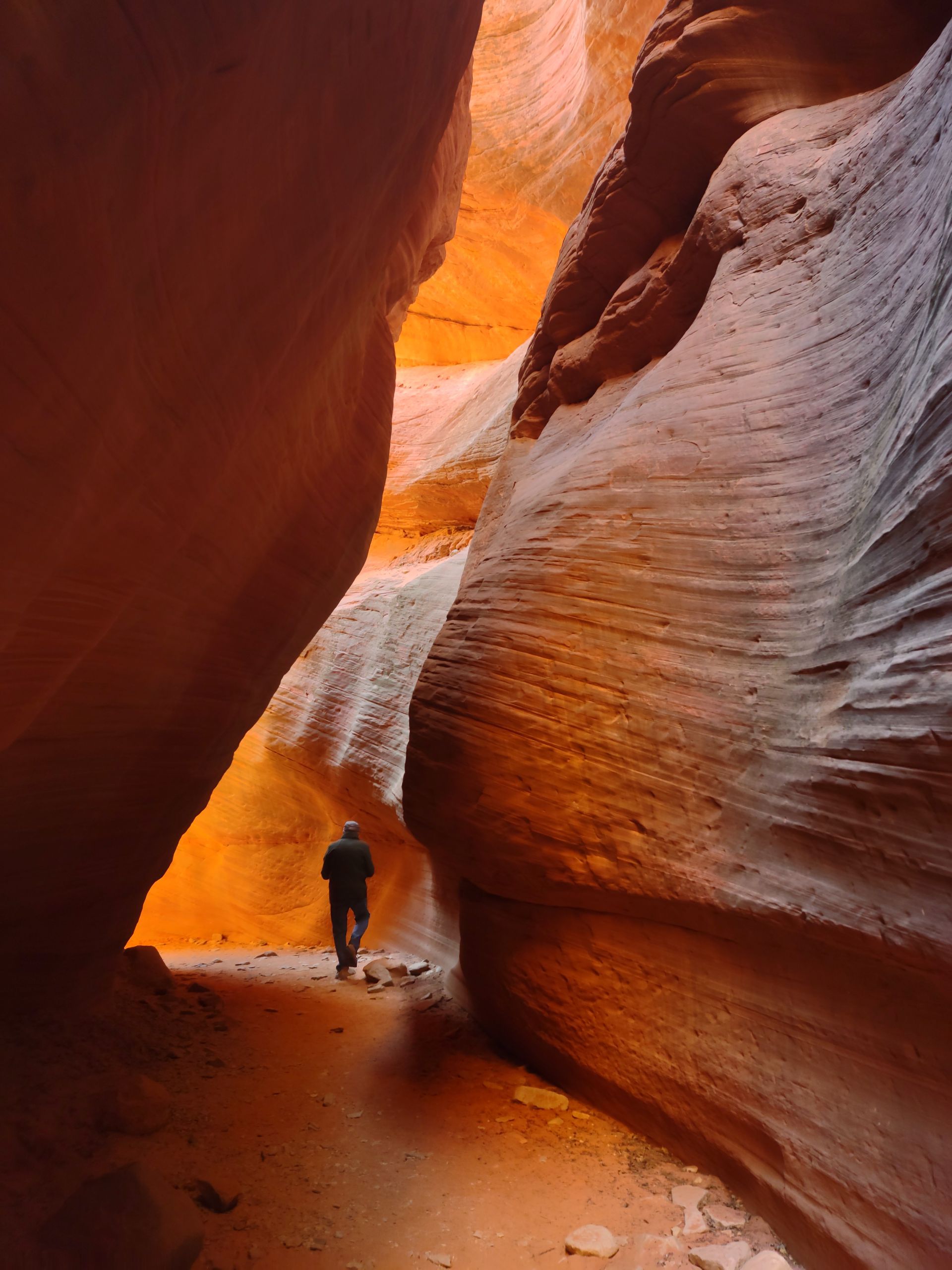 Person walking through a narrow slot canyon with orange and tan rock walls. Light streams from the canyon's opening.