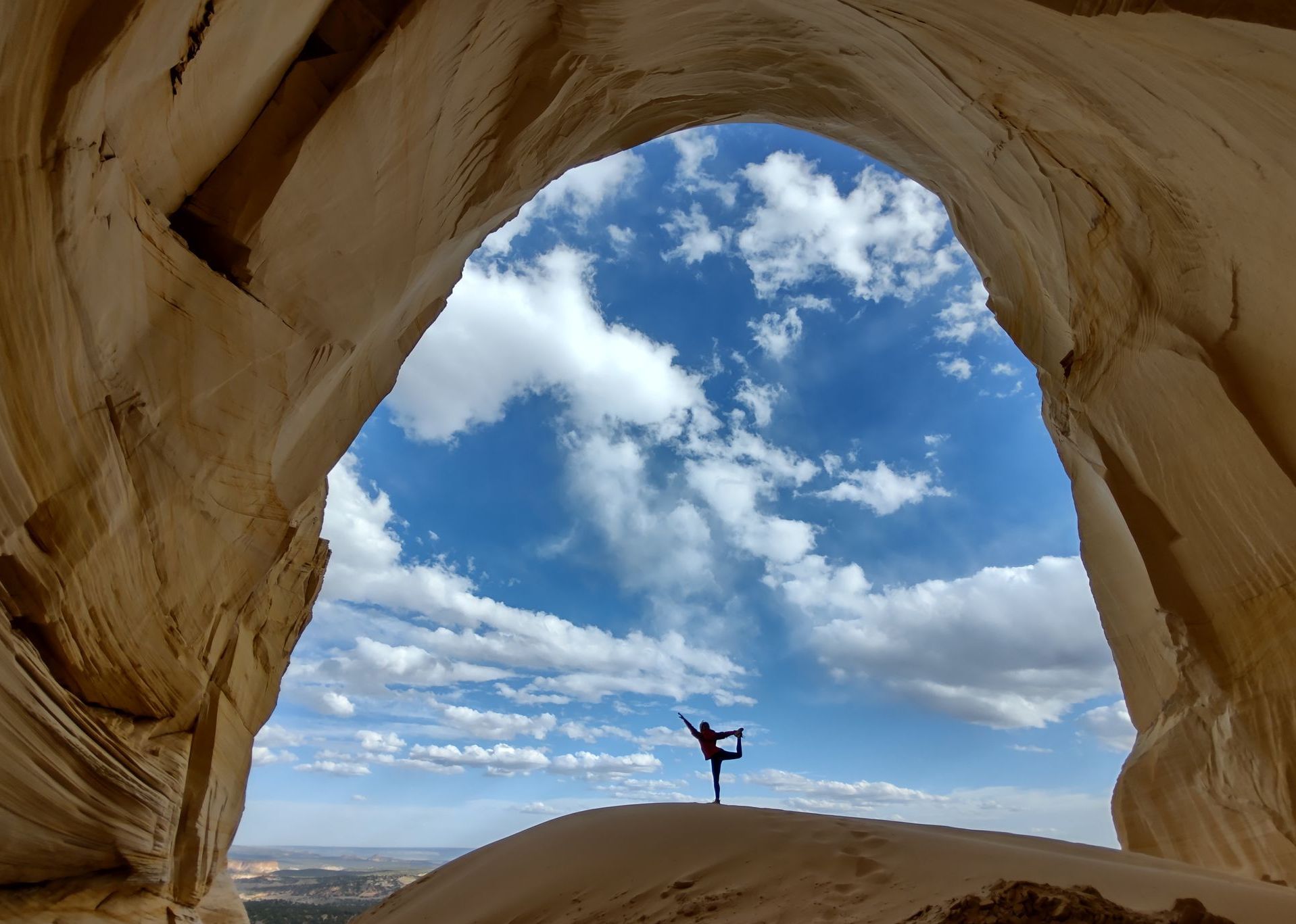 Woman balances on one leg, arms outstretched, against a backdrop of blue sky and clouds, viewed through a sandstone arch.
