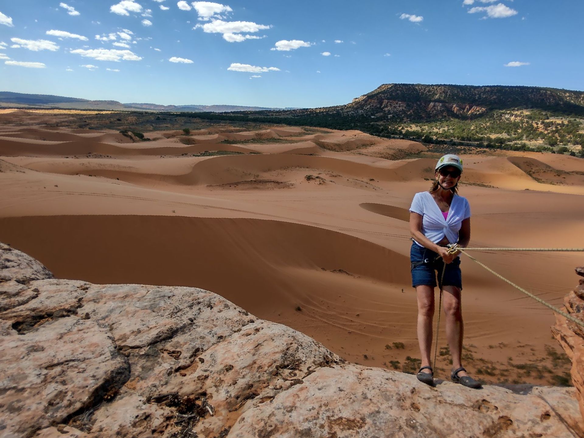 Woman on rock cliff with sand dunes in the background. She is holding a rope. Blue sky with clouds.