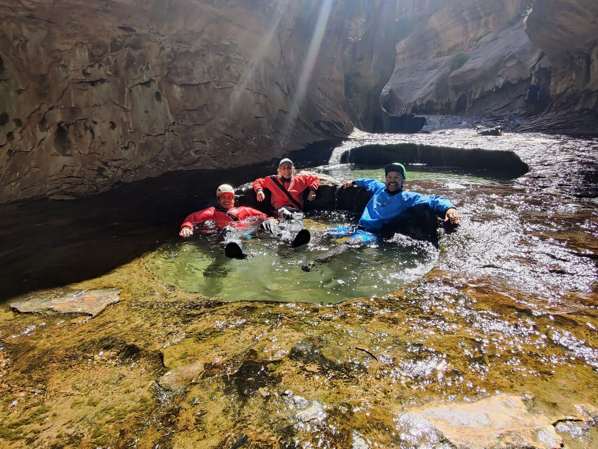 Three people in wetsuits relax in a natural pool of clear water within a canyon.