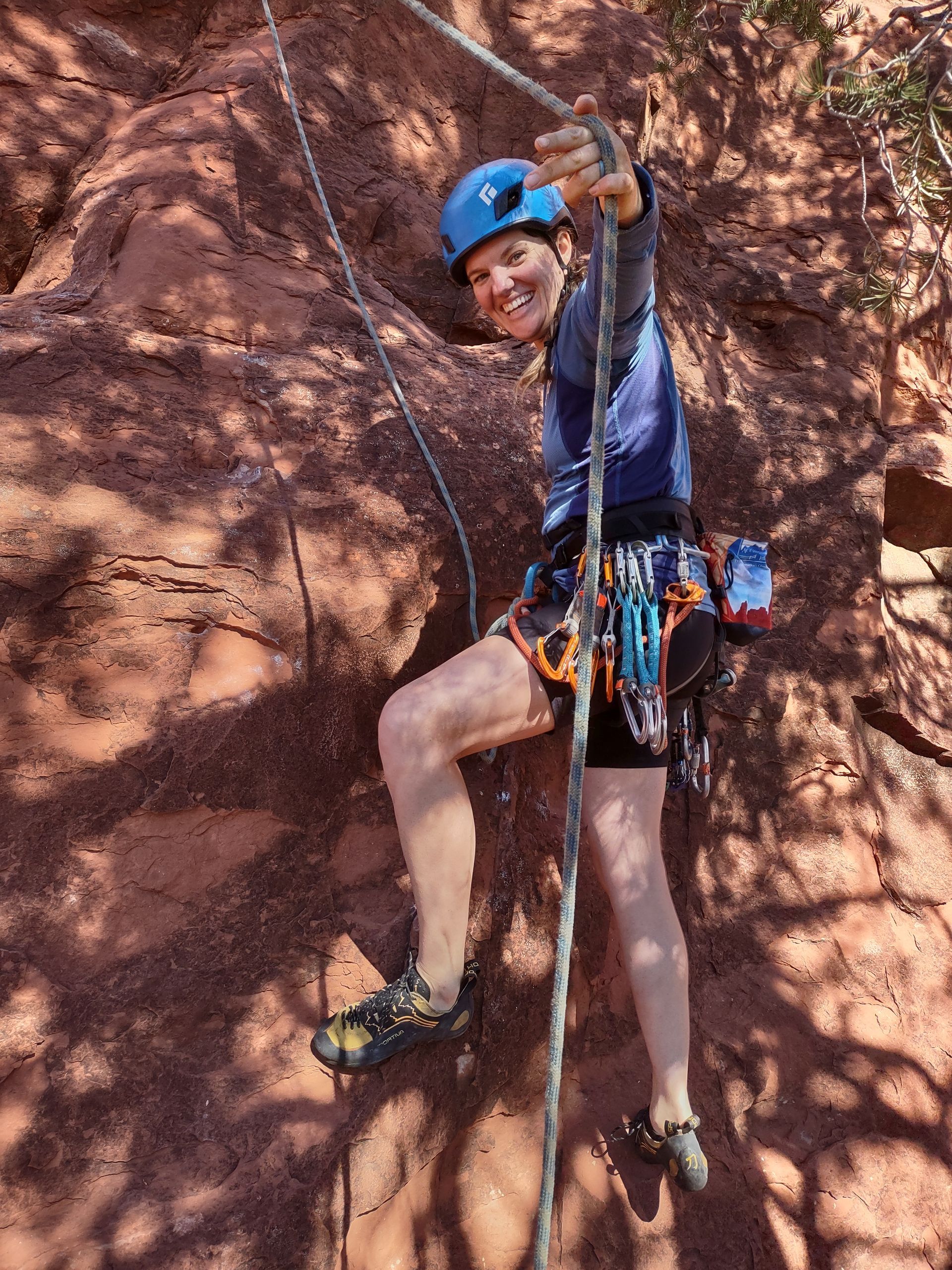 A person rock climbing, smiling, with a helmet, harnesses, and gear, attached to a rope on a red rock wall.