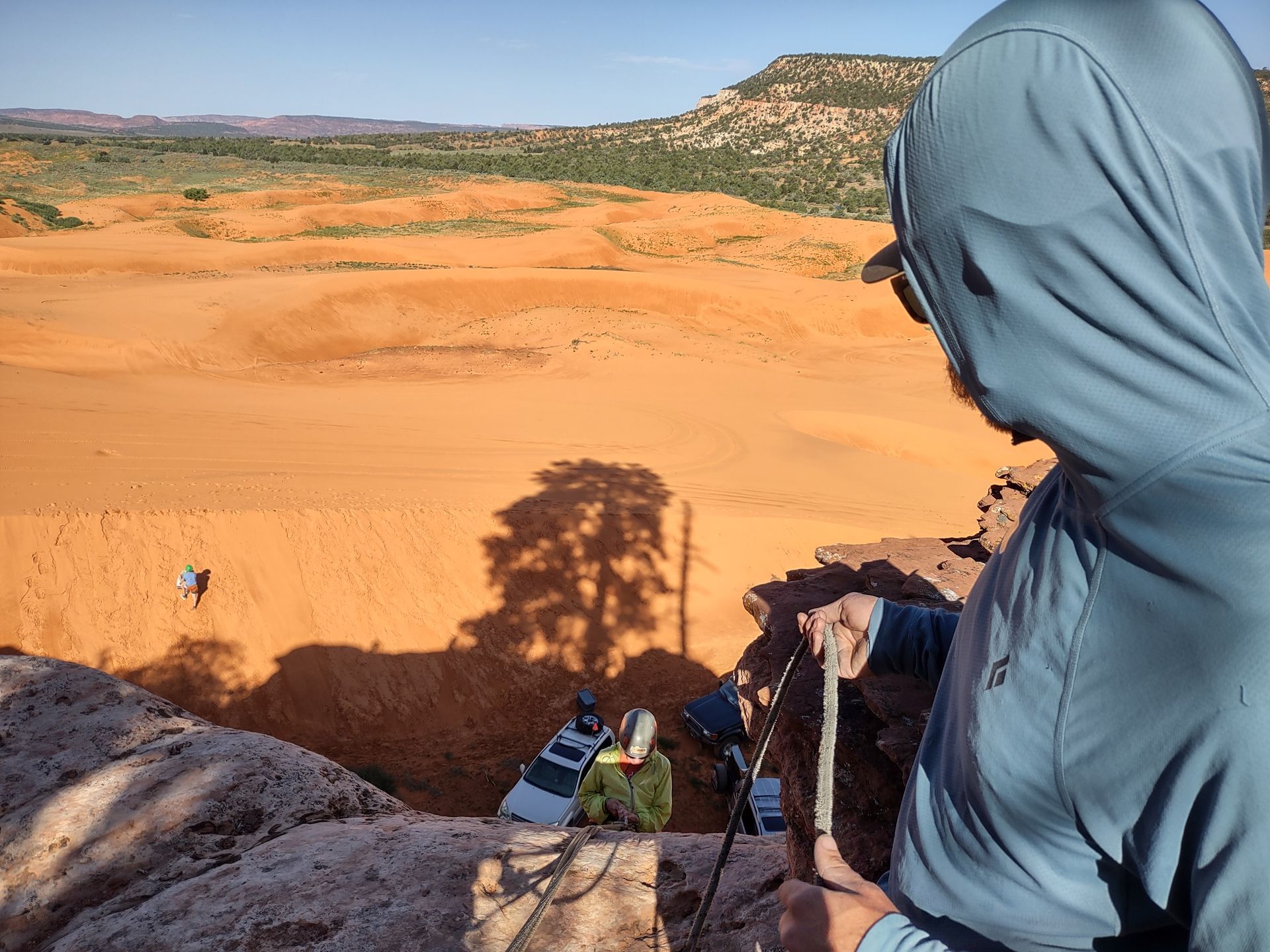 Person in blue hoodie, looking down at people in a desert landscape. Shadows of a tree and people are visible.