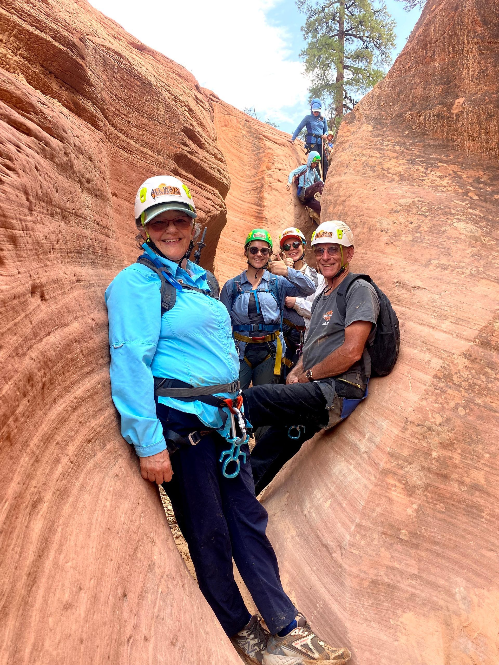 Group of people canyoneering in a narrow red rock canyon. Two in foreground smiling, wearing helmets and harnesses.
