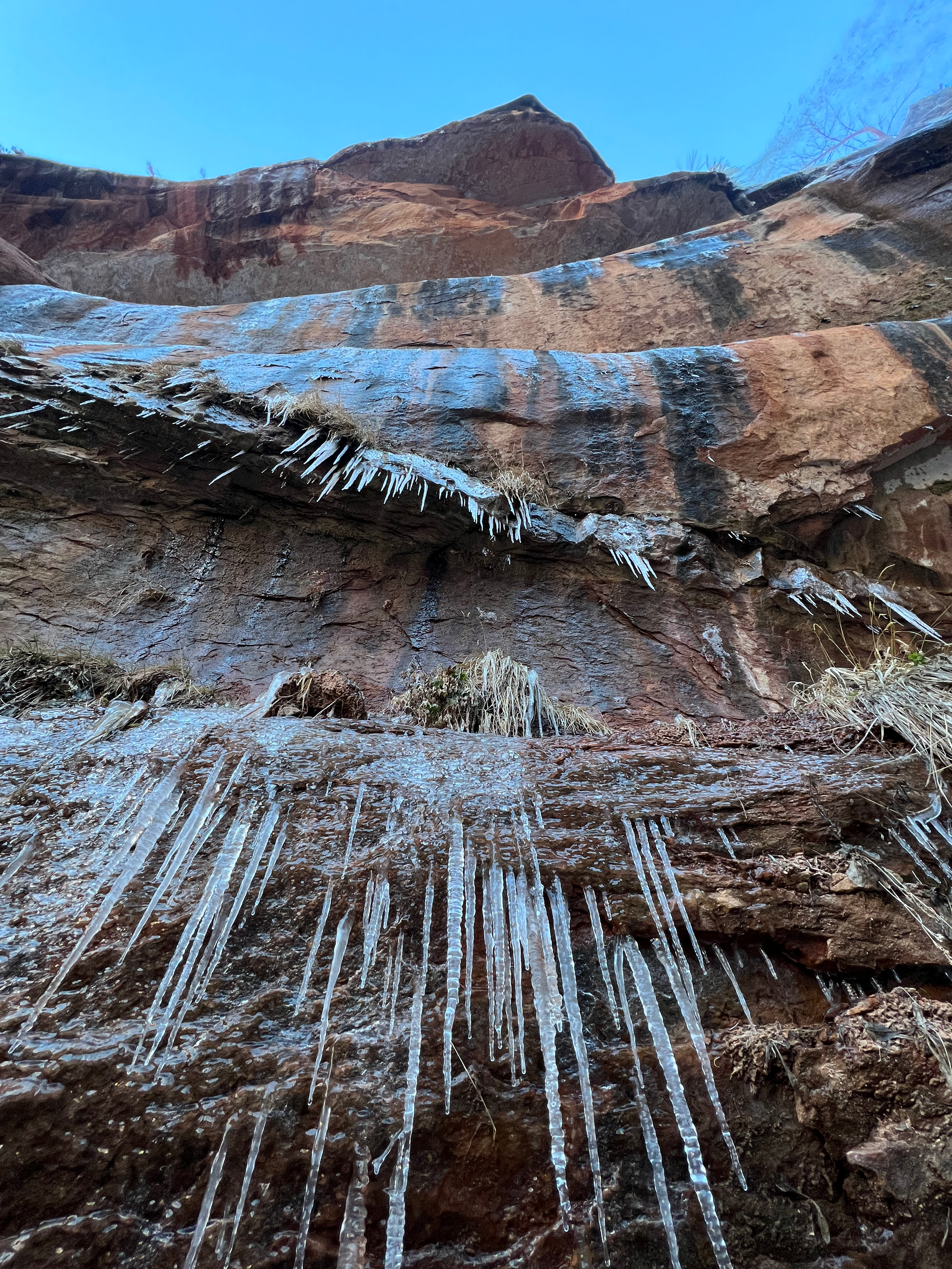 Icicles hang from a reddish-brown rock face against a blue sky.