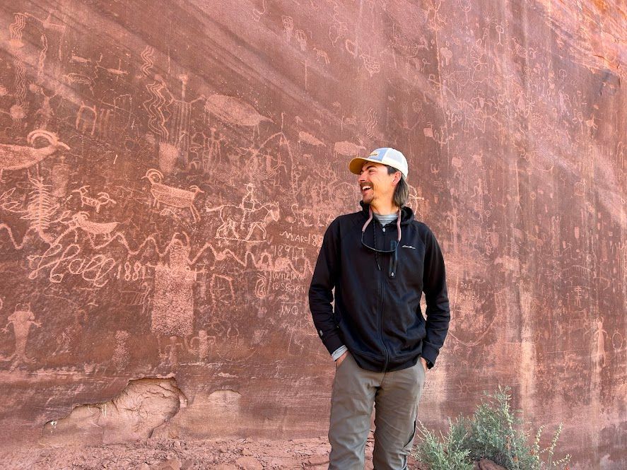Man smiles at ancient rock art on a red sandstone wall. He wears a cap, black hoodie, tan pants.