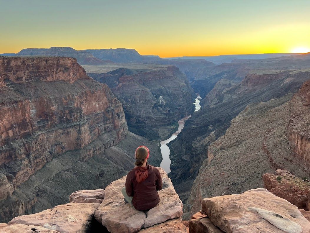 Person seated, facing scenic canyon vista at sunset, with river below.