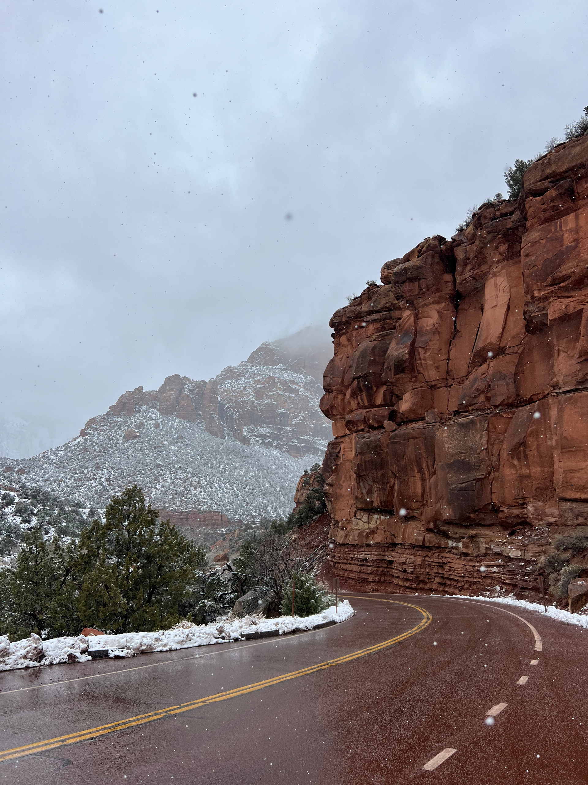Snowy road curves past red rock cliffs, overcast sky.