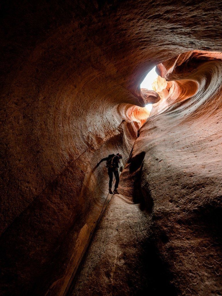 Person rappelling down a narrow, reddish-brown slot canyon. Sunlight streams from the opening above.