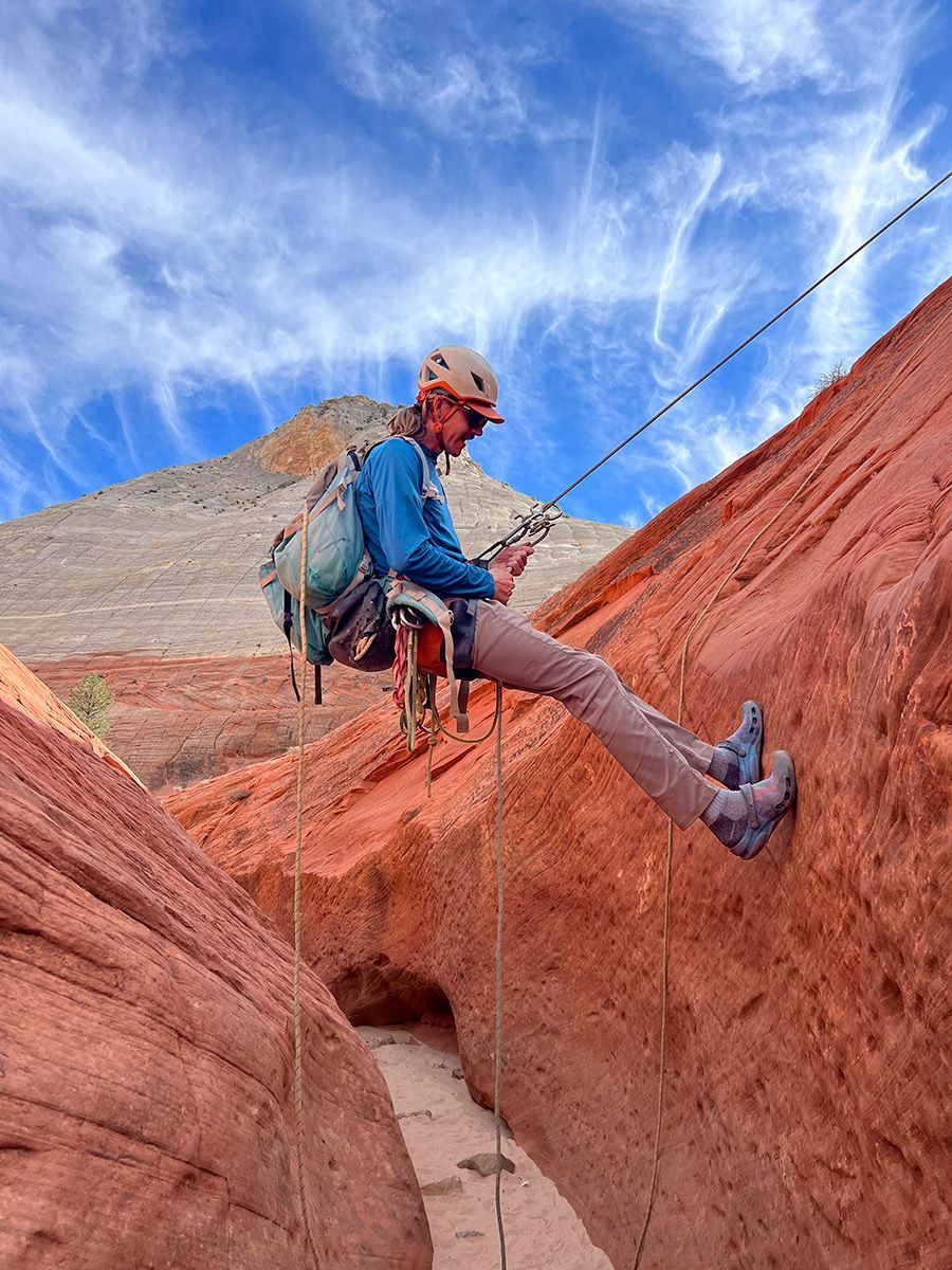Person rappelling down a red rock canyon with a blue sky above.