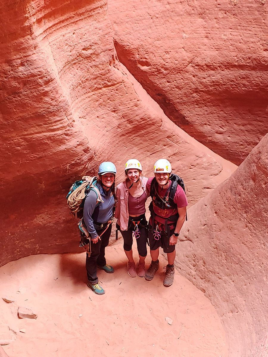 Three people in helmets and harnesses stand in a narrow, red-rock canyon.
