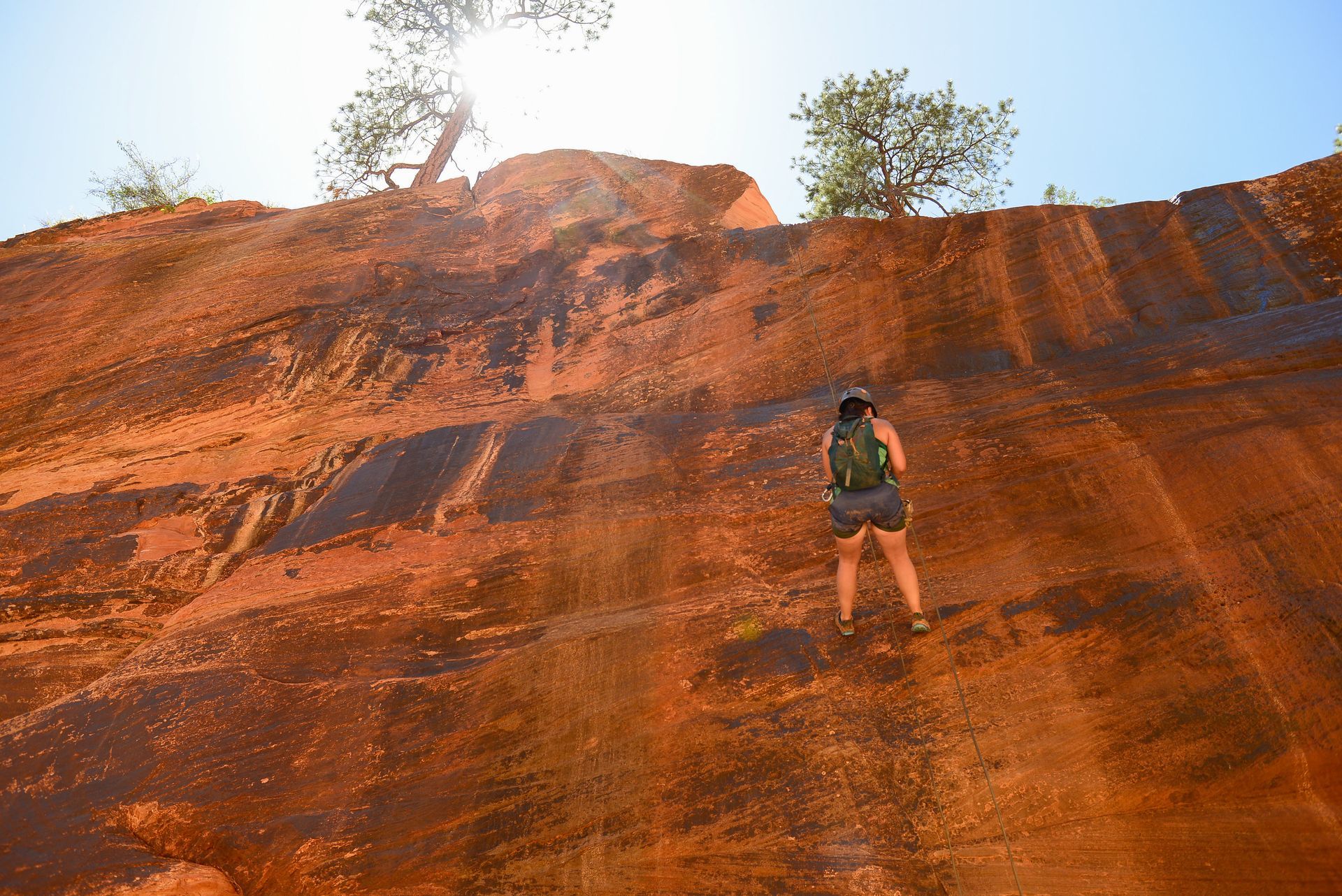 Person wearing backpack climbs red rock formation, sunny outdoor setting.