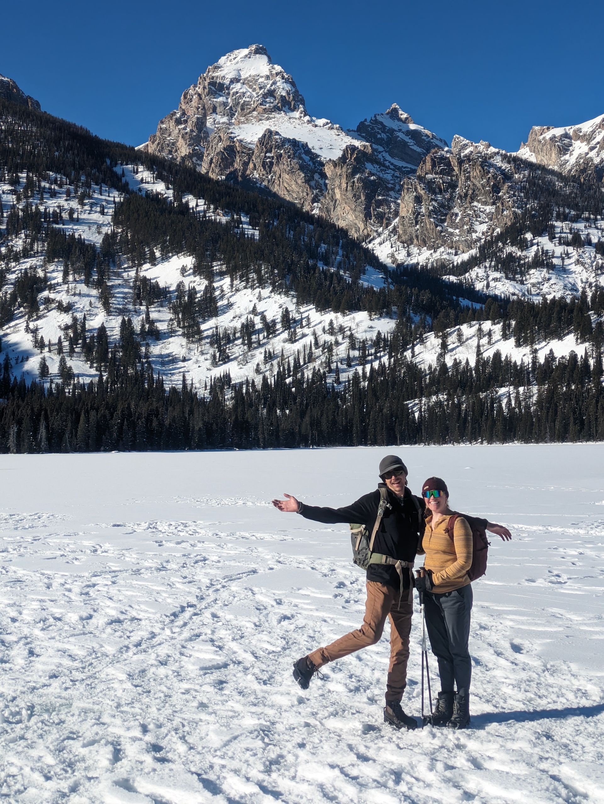 Two people in winter gear pose on a snow-covered landscape with mountains in the background. Sunny day.