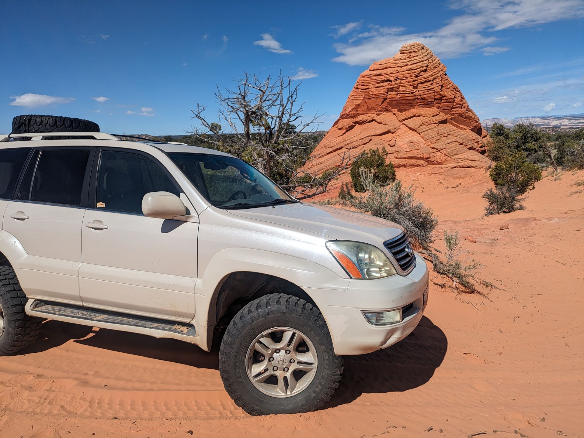A white SUV parked on red sand with a rock formation in the background under a blue sky.