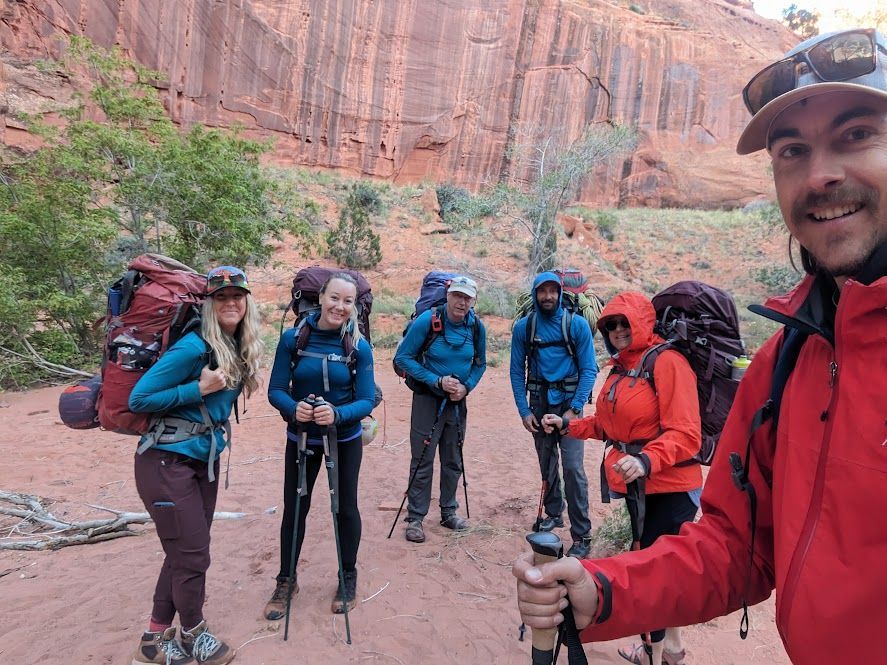 Group of hikers with backpacks, smiles, and hiking poles pose in a red rock canyon.