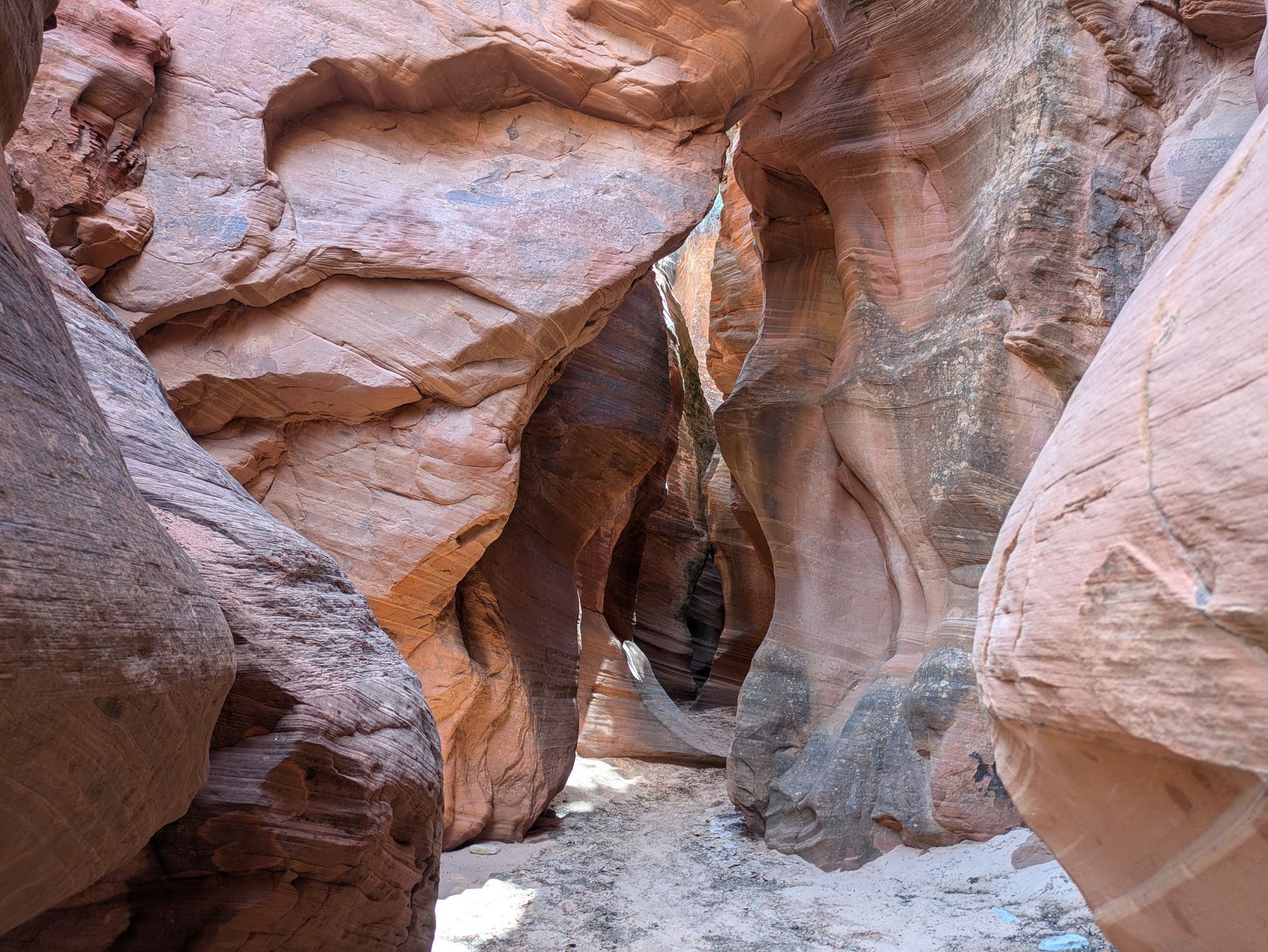 Narrow canyon with orange and brown sandstone walls, a sandy floor, and sunlight filtering in.