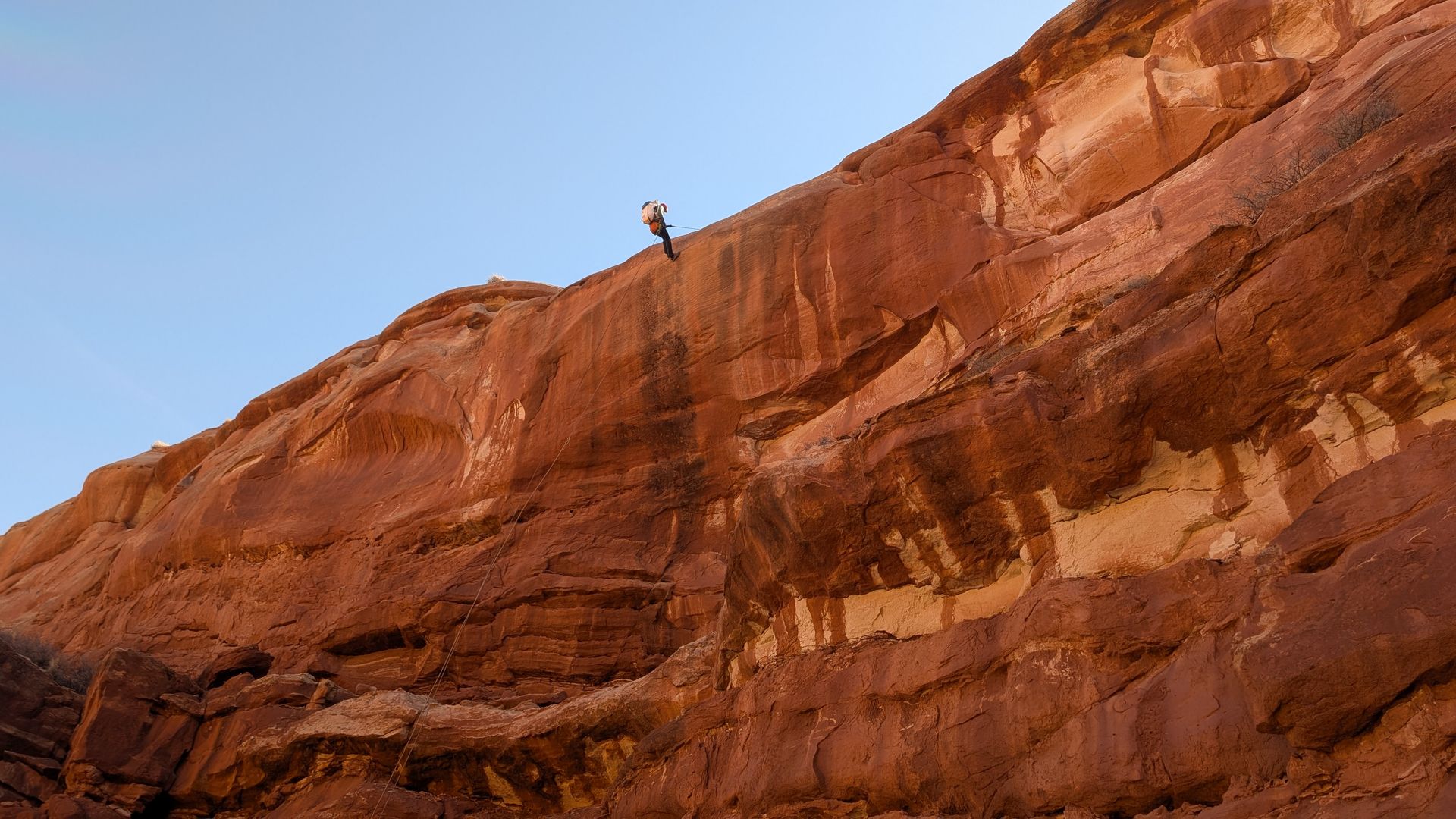 Rock climber rappelling down a tall, red sandstone cliff, with clear blue sky above.