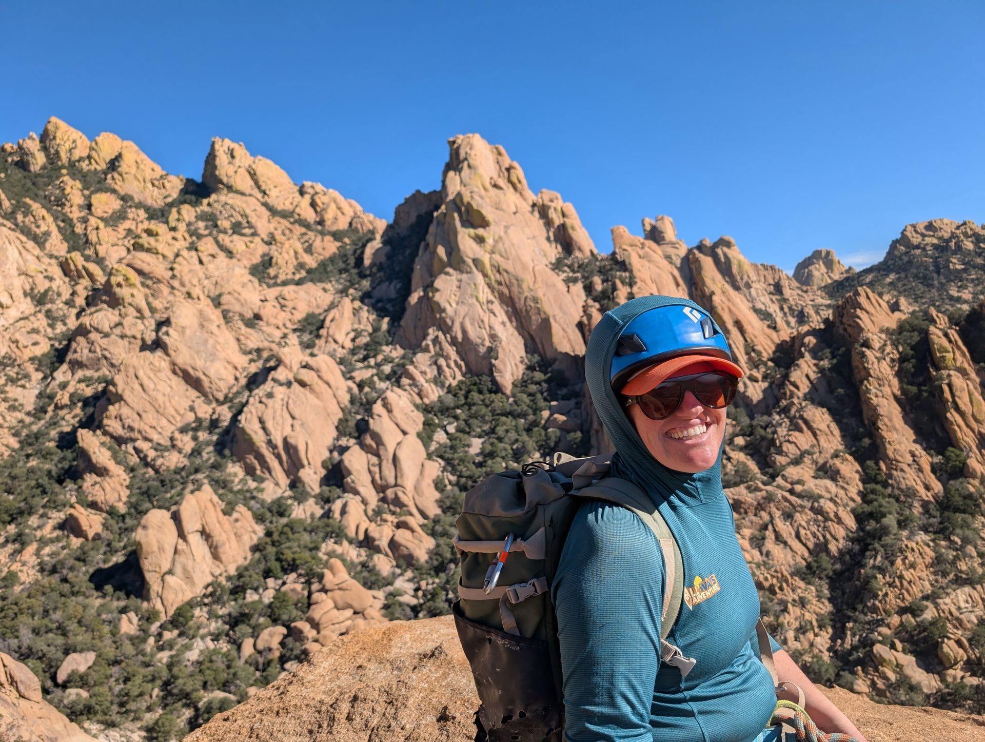 Woman in climbing gear smiles, rock formations in the background. Sunny day.