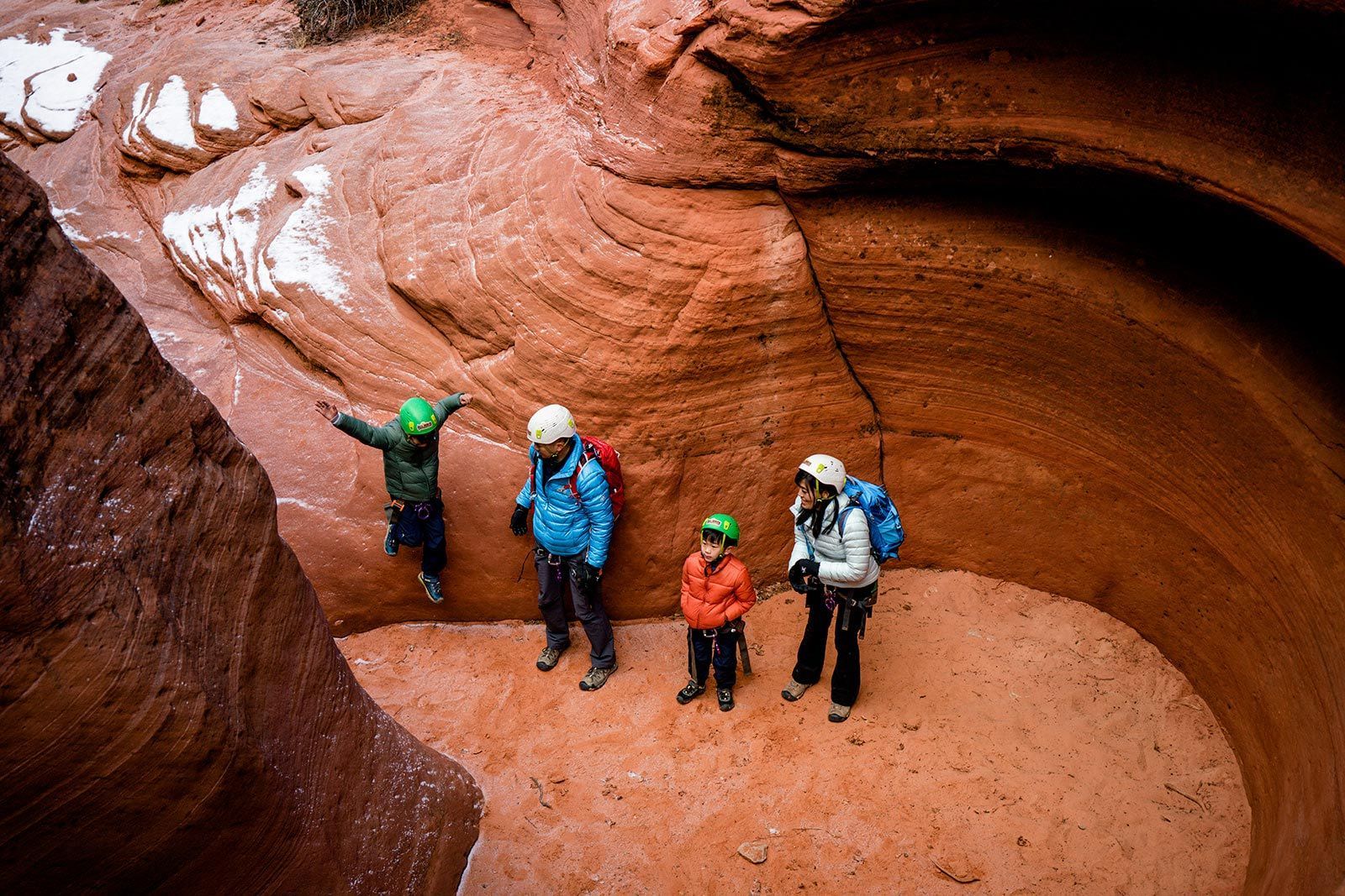 Four people in a red rock canyon, wearing backpacks and looking up. One child is jumping.