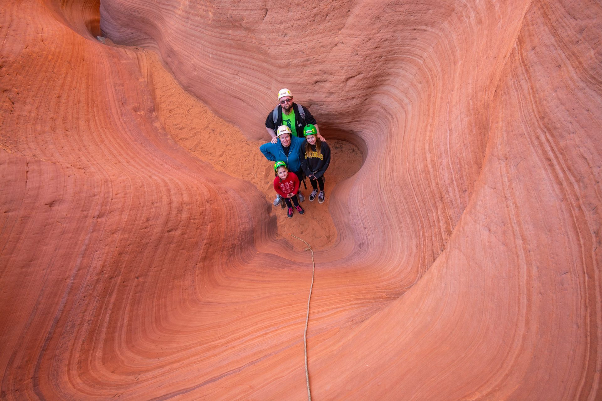 Four people wearing helmets and outdoor gear stand at the bottom of a spiraling, orange sandstone slot canyon.