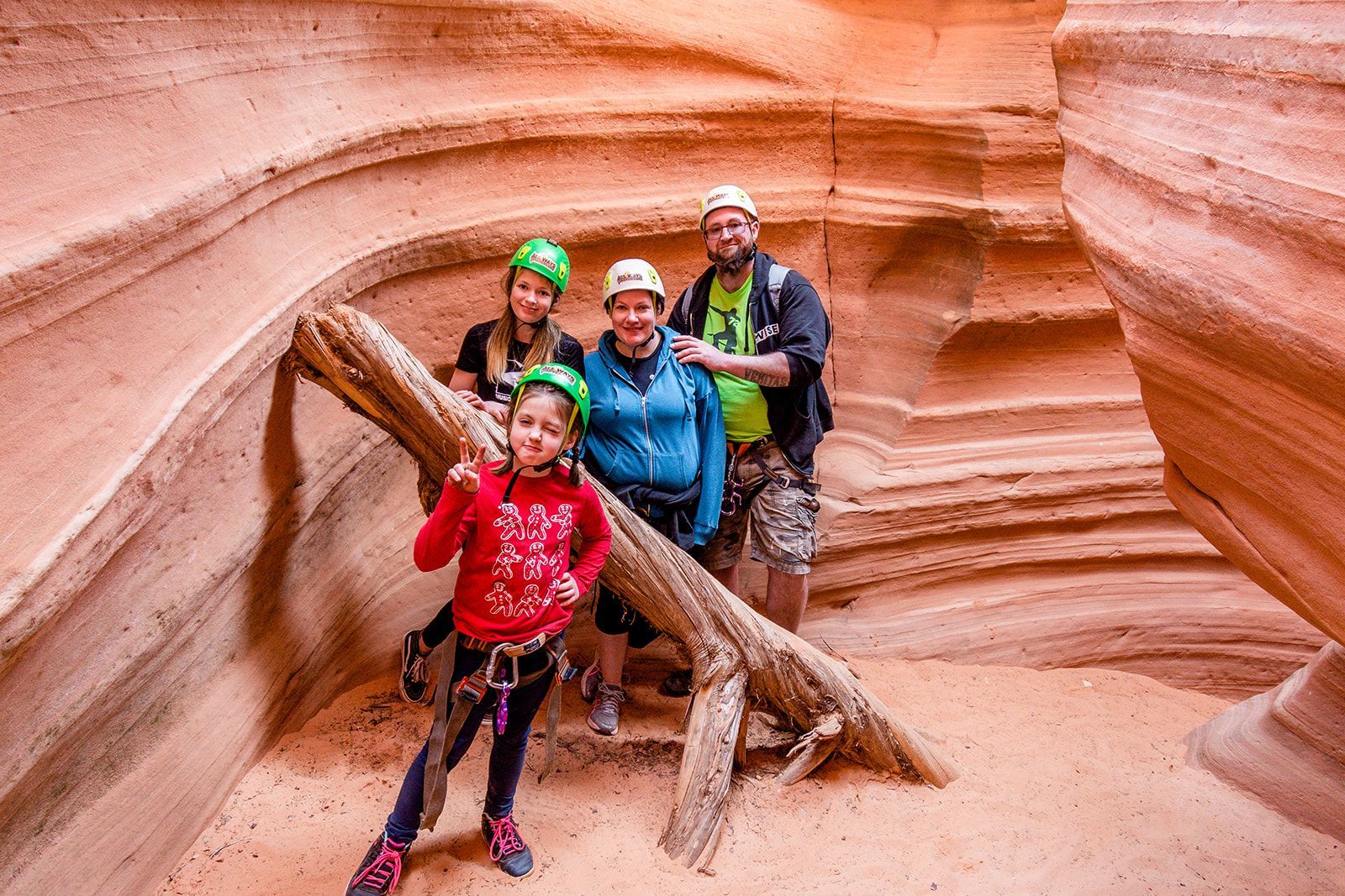 Family of four with helmets and harnesses in a narrow, orange sandstone canyon. They smile at the camera near a fallen log.
