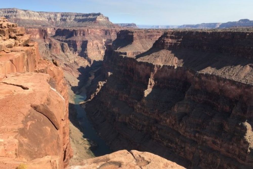 Grand Canyon vista: Red rock formations, canyon walls, and a river below under a clear blue sky.