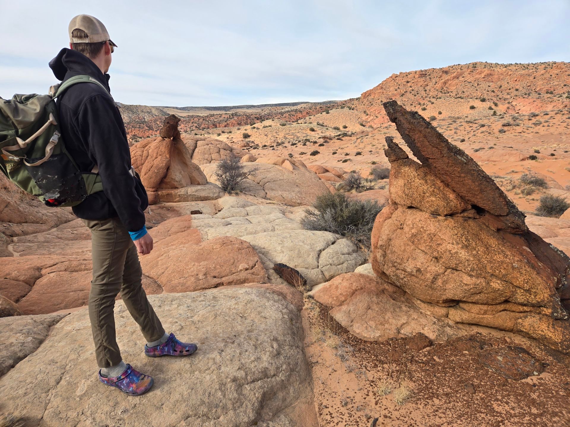 Person hiking in a desert landscape, looking at rock formations. They wear a hat, backpack, and hiking shoes.